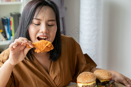 包子小吃饥饿的超重女持有炸鸡木板上的汉堡包和桌披萨在家工作期间体重增加问题百食不厌症BED概念成人图片
