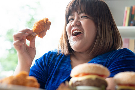 盘子加大尺码一顿饭饥饿的超重女持有炸鸡木板上的汉堡包和桌披萨在家工作期间体重增加问题百食不厌症BED概念图片
