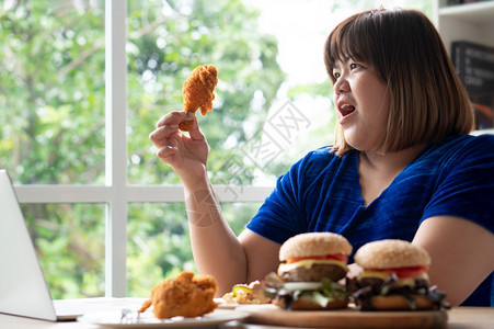 垃圾美味的饥饿超重女持有炸鸡木板上的汉堡包和桌披萨在家工作期间体重增加问题百食不厌症BED概念健康图片