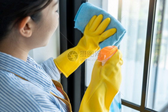 蓝色的家庭主妇身戴明亮黄色手套的年轻亚洲妇女洗脸或打扫窗户玻璃上用蓝色抹布擦灰和洗涤尘土污渍她公寓的洗涤剂服务图片