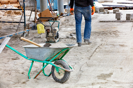 路面基础建筑工地用一个轮式手推车和一个铲子来运送沙以便在工人和模糊的振动板上铺设在工人背景下铺设面板的石以及用沙子和铲在施工地铺图片