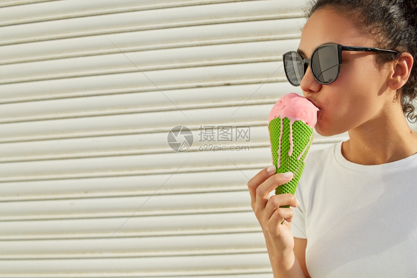 可爱的美国人穿着白色T恤和轻牛仔裤的年美籍黑人女孩在日光照耀时用冰淇淋抵挡着一堵轻墙吃冰淇淋热的图片