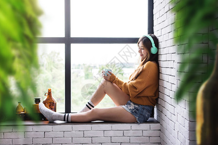 坐年轻女在靠近窗户的CozyHouse家享受阳光和热咖啡的早与清晨请享用时尚图片