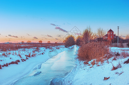 水平的气候乡村冬季夜晚雪田下有冰覆盖的河日落时有一片粉红下的远屋和蓝天白日之下有蓝色天空的遥远房屋白雪皑图片