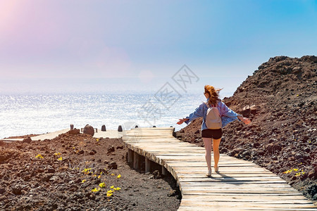 微笑散步欧洲背着包的年轻女子从岩石岸边的木平台上走到大海在阳光明媚的日子里在海边快乐的女人明亮蓝天特内里费岛的火山岩年轻女子背着图片