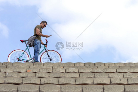 特伦迪男人坐在固定自行车上时在户外使用智能手机保持太阳镜城市图片