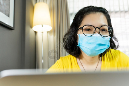 人亚洲快乐女在隔离期间使用笔记本电脑戴眼镜黄色衬衫中孤立的年轻女青穿着蓝色外科面具在流行Covid19科罗纳健康预防保护等流行病图片