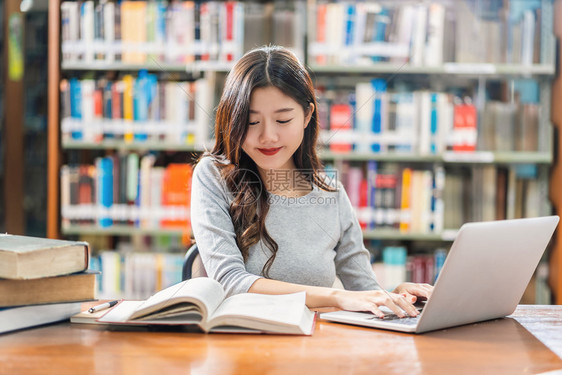 上市做功课和在大学图书馆或使用技术笔记本电脑的亚洲青年学生或具有各种书籍和文具架背景的各种群落返回学校从事家庭作业和使用技术笔记图片