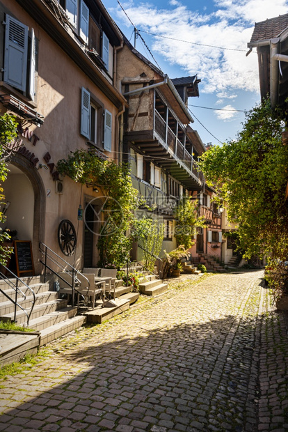 生产法国阿尔萨斯州Eguisheim中世纪镇沿一条狭小街道上带有可腐石和绿色植物的多彩有半形房屋木结构歪图片