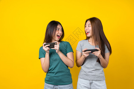 使用智能手机以孤立的黄色背景生活方式和业余爱好概念玩游戏的两幸福亚洲笑着年轻女子游戏家生活风格与休闲愉快曼谷年轻的图片