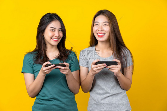 游戏玩家时髦的使用智能手机以孤立的黄色背景生活方式和业余爱好概念玩游戏的两幸福亚洲笑着年轻女子游戏家生活风格与休闲女图片