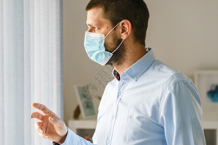 病人非典站在窗边的caucasian男子旁观他们身着医疗保护面罩在家中隔离或办公室看外衣时工作防止细菌流行传染病同时要防身穿衬衫图片
