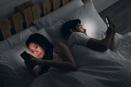 网络微笑互联在床上的年轻夫妇冲突快乐的笑着女人将她转回男身边阅读手机上的信息试图偷看屏幕变相和不忠的概念图片