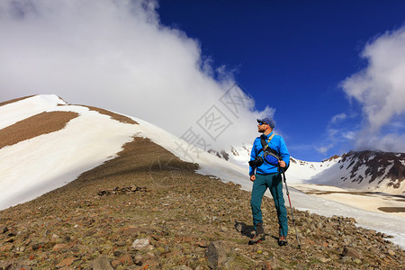 多云的探索登山手拿着一根拐杖的旅游摄影师站在山顶上一个雪峰高山坡上并计划在一个明亮蓝色和阴云的天空摄影师背景下攀登旅游摄影师检查图片