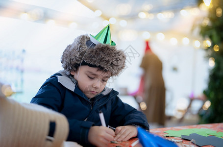 坐着花粉笔绘画或写纸彩模糊明亮的浅光背景儿童在生日派对上玩乐儿童在圣诞节或新年日从事活动等有政党帽子的可爱男孩写作或者图片