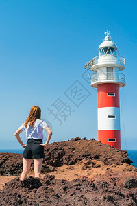 年轻女子站在岩石海岸的灯塔前女孩靠近火山岩上的法尔阳光明媚的日子亮蓝天海岸上灯塔女孩靠近法尔在火山岩湛蓝的天空海岸上灯塔自然晴天图片