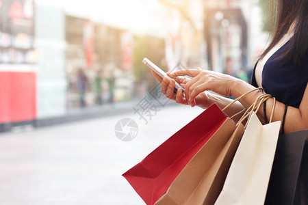 买方天妇女站在商场背景上时使用智能手机和持有购物袋的妇女以及新图片