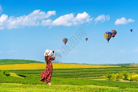 日出旅游蜜月妇女站在绿草上用智能手机给热空气球拍照她们站立在绿色草地上用智能手机照相图片