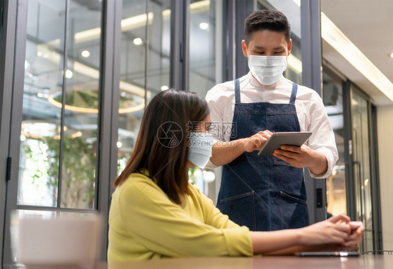 桌子黄色的服务员佩戴保护面罩接受亚洲女客户在现代咖啡馆或餐中带有数字石片的亚裔女客户订单在公共场合有新的正常生活和方式的人亚洲图片