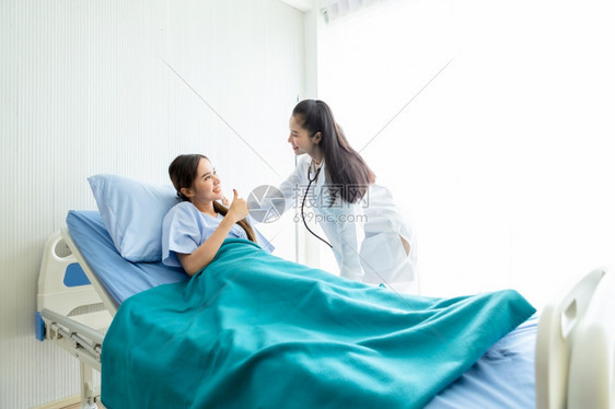 专家亚洲年轻女病人在床上展示拇指与微笑脸的相容对医院背景中的亚洲年轻女医生来说考试大拇指图片