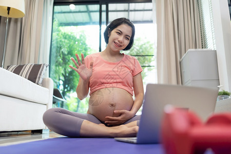 年轻的孩子沟通亚裔孕妇在家中用笔记本电脑进行视频聊天时在家中微笑的年轻母亲说话并用笔记本向家人挥手女在怀孕期间做瑜伽练习图片