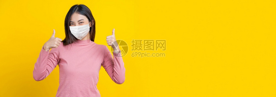 身戴面具和大拇指的亚洲微笑妇女将行动作为毕业横幅或展示孤立黄色背景科罗纳流行可知19年爆发旗帜和美与共体保护乐趣室内的工作图片