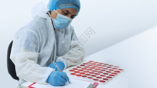 戴着面具和蓝色手套的医生在一张白纸上写字旁边是一组白色圆形容器中的红色液体临床样本圆形白色容器中红液体的临床样本写作生物学坐着图片