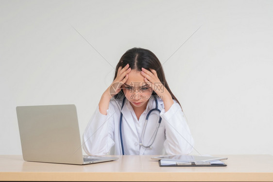 脸年轻的亚洲女医生坐在办公桌旁由于过度工作精神压力疲劳的概念和医生责任她看起来不快乐和疲倦处理沮丧的病情桌子治疗图片