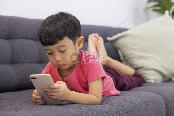 微笑的小男孩看着坐在客厅舒适沙发上的触摸板享受在数字智能手机上玩游戏的乐趣利用有的应程序快乐小孩子笑声无线的家在图片