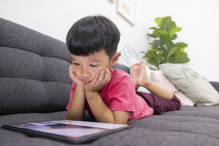 微笑的小男孩看着在客厅舒适沙发上的触摸板享受在数字平板电脑上玩游戏的乐趣利用有的应程序来玩乐小孩子游戏活的技术保持图片