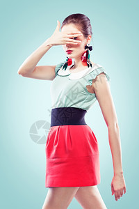 小型的Retro风格的年轻女子用一只手将脸闭上但看她的手指中间照相机穿着大漂亮的耳环蓝色背景的耳环短裙收入图片