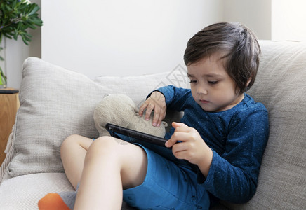卡通片年轻的亚洲人可爱小男孩在平板上玩游戏儿童在数字小盘上看卡通Kid坐在沙发上好奇的脸在触摸垫上玩游戏图片
