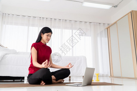坐着亚洲人关心健康的亚洲年轻妇女在卧室里进行瑜伽锻炼在家里用笔记本电脑在线学习运动和放松概念技术促进新正常生活方式的创新与放松概图片