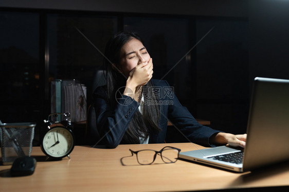 尽管黑暗的亚洲女商人坐在打哈手盖着她的嘴笔记本电脑在桌子上而这位年轻女晚上在办公室加班沮丧的图片