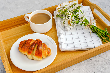 牛角包咖啡加奶新鲜出炉的羊角面包方格餐巾纸和甘菊花放在灰色石桌上的木托盘制舒适图片