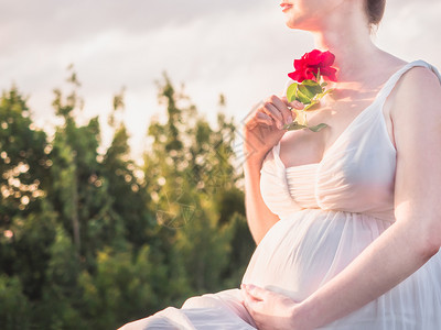 公园生活年轻妈穿着时尚白色的衣服站在清澈的天空和阳光下手里拿着一朵美丽的红玫瑰花幸福的家庭和母亲年轻妈穿着时装白色的礼服一种图片