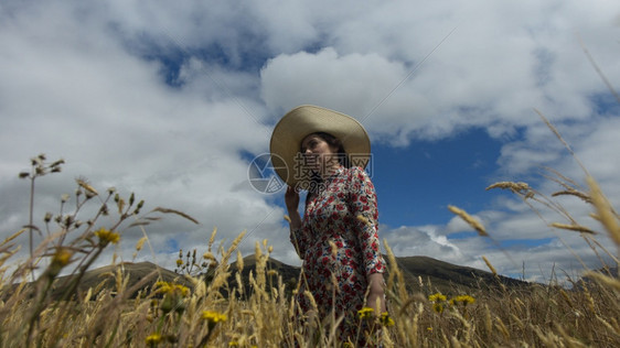 时尚乡村的穿着花裙和长帽的快乐年轻女子在阴天中走麦田间背景是蓝天和山脉多云的麦田女孩图片