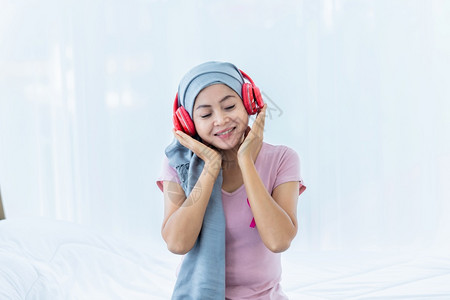 微笑一位患有乳癌的亚裔女患者快乐她带着粉红色丝在耳机上戴头巾正在听音乐治疗后化就坐在床上房子里的卧室家屋图片