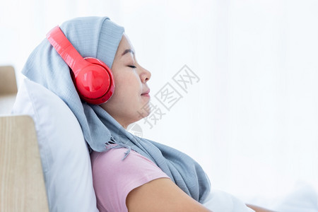 屋幸存者一位患有乳癌的亚裔女患者快乐她带着粉红色丝在耳机上戴头巾正在听音乐治疗后化就坐在床上房子里的卧室放松图片