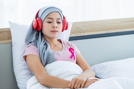亚洲人高级的收音机一位患有乳癌的亚裔女患者快乐她带着粉红色丝在耳机上戴头巾正在听音乐治疗后化就坐在床上房子里的卧室图片