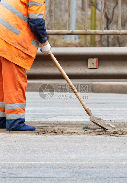 维护路面工作一名穿橙色反射制服的清洁工在金属保险杠垂直图象下从两条小道之间的公路上流出沙子复制空间清洁工用铲子将金属保险杠下路道图片