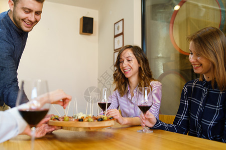 一顿饭坐在桌边的两名女天主教孩拿着红酒杯而一名年轻男子正在家里或餐馆为一天在家中微笑的开胃小吃朋友服务开胃酒成人图片