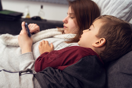 童年睡在沙发上或床玩游戏手机智电话或在隔离期间晚上家用视频打字时Smms发短信的幼小母亲和儿子在隔绝期间家庭可爱的图片