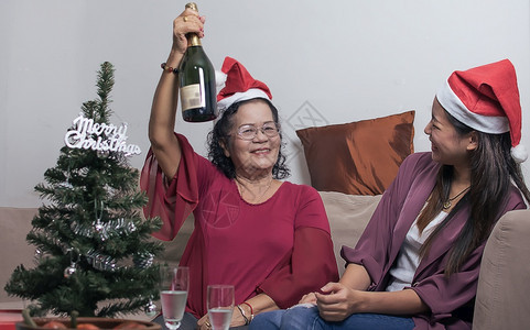 活的亚洲人两个60岁的老母亲和女儿戴着圣塔帽在坐家中客厅时一起喝酒头寸模糊的树前景瓶装圣诞或新年庆典概念圣诞节或新年庆典概念老的图片