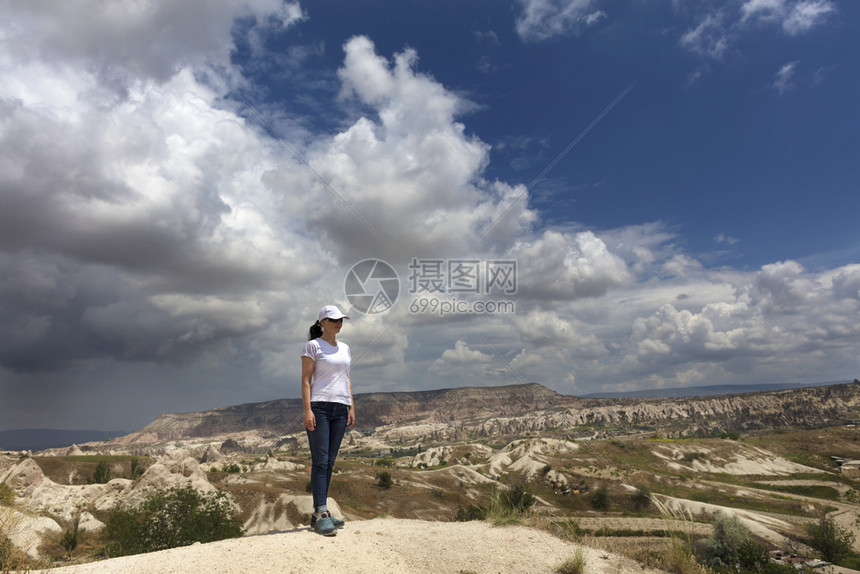 站立一个戴着白色帽子和T恤蓝色牛仔裤的年轻女孩站在卡帕多西亚的一块岩石上在多云的天空和山景背下望向远方一个年轻的女孩站在卡帕多西图片
