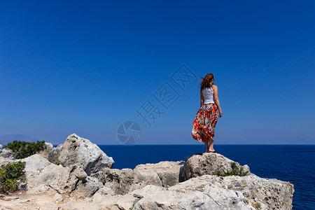 身穿多彩裙子和白色上衣的年轻女子站在悬崖边缘看着平淡蓝色海面边景岩石缘很多空气有模仿间的图像女士站在石头悬崖的边缘望向海平面的距图片