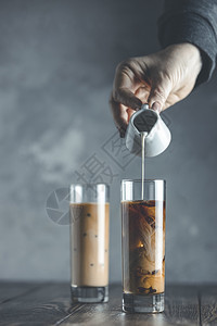 目的灰色新鲜妇女用手把自制酸奶油从小罐子倒到玻璃煮冷咖啡和冰的夏季饮料放在黑木桌和灰色背景及复制空间上图片