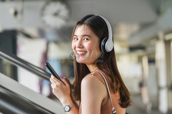 车站小时年轻亚洲女乘客在大城市通勤和交上旅行时佩戴耳机和使用智能移动电话聆听音乐并走在地铁站的楼梯上如乘坐大城市往返车和交通疏远图片