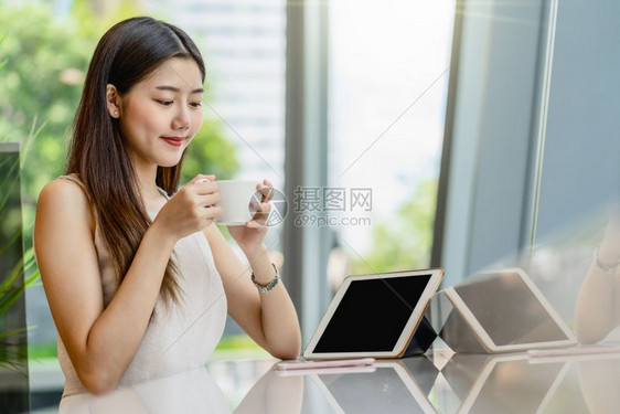 亚洲年轻女在现代咖啡店或工作场所与窗户镜子日本朝韩生活方式和日常企业家概念旁边的玻璃窗外在现代咖啡店或合用办公空间内喝着咖啡和看图片