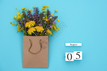 自然开花平坦的Wooden立方体8月5日历和蓝背景的手工艺品包中富彩的生化花朵蓝色背景的GreetingcardFlatLay概图片